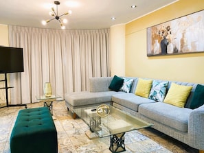 Una sala de estar con un sofá modular, una mesa de centro de vidrio, un reposapiés color turquesa, un televisor de pantalla plana y un cuadro en la pared.