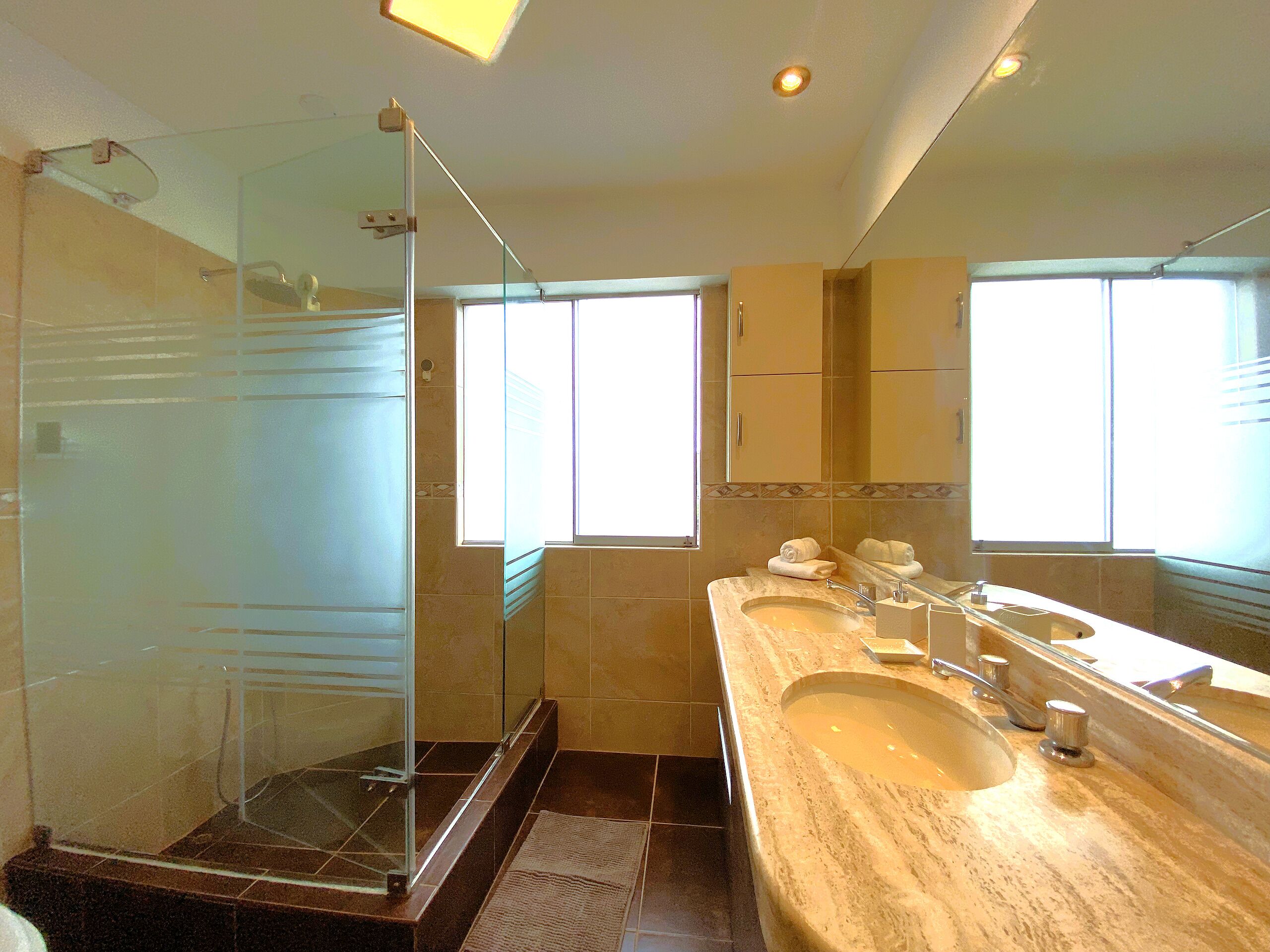 A bathroom with a glass shower enclosure, a sink with a large mirror, and a window providing natural light.