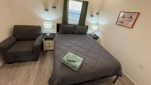 Un dormitorio con cama, una silla, mesitas de noche, una ventana con cortinas y un cuadro en la pared.