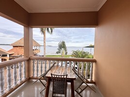 Un balcón con una mesa y sillas de madera, barandal blanco y vista a un cuerpo de agua con edificios y palmeras al fondo.