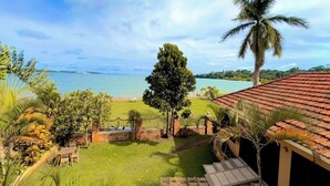 A view of a lush garden with a palm tree, a wooden bench, and a pathway leading to a beach.