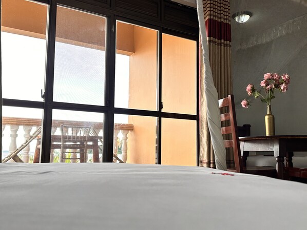A hotel room with a large window, a bed, a chair, a small table, and a vase with flowers. - Sienna Beach Hotel - Entebbe (Entebbe)