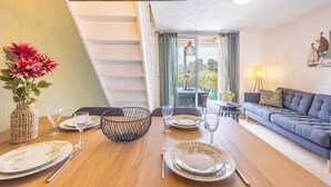 A dining area with a wooden table set for four, a wicker vase with flowers, and a blue sofa in the background. - Clos Fontaine House with garden, pool (Valras-Plage)