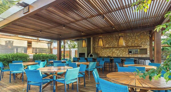 An outdoor patio with wooden tables and blue chairs, a stone wall, and a bar area. - Apt in Condo w/ Pool in Barra Grande Vgd0001 (Maraú)