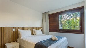 Quarto com cama, mesa de cabeceira, janela com vista para palmeiras e cabeceira de madeira.