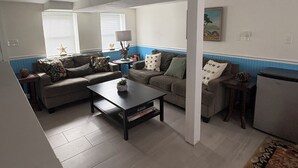 Sala de estar com dois sofás, uma mesa de centro e uma mesinha lateral. Há uma janela com persianas e um quadro na parede.