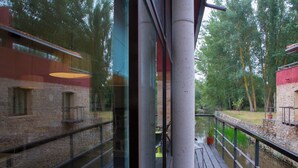 Een houten terras met een reling, met uitzicht op een rivier, stenen gebouwen en bomen op de achtergrond.