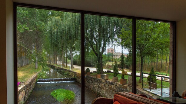 Een kamer met een groot raam dat uitkijkt op een tuin met een waterpartij en bomen.