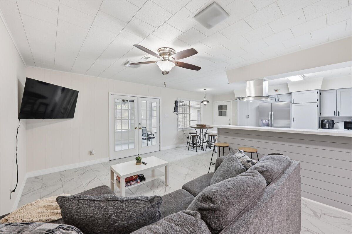A modern living room with a grey sofa, a flat-screen TV mounted on the wall, a ceiling fan, and a kitchen area with white cabinets and a stainless steel refrigerator.