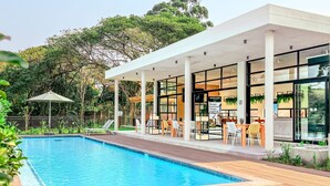 A modern pool area with a wooden deck, outdoor seating, and a covered patio with glass walls.