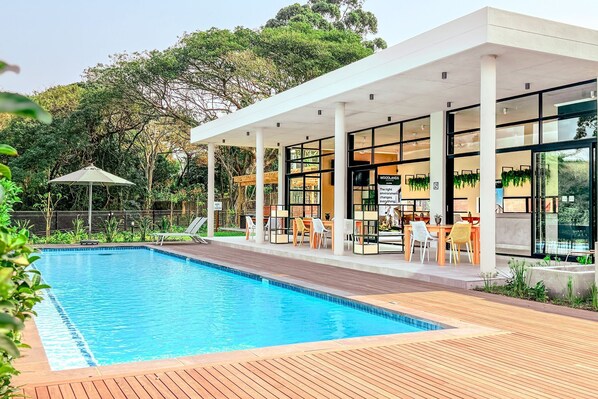 Un área de piscina moderna con cubierta de madera, asientos exteriores y un patio cubierto con paredes de vidrio.