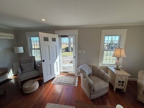 A living room with a wooden floor, a grey armchair, a beige armchair, a small wooden side table with a lamp, and a white door leading outside. - Waterfront Farm Cottage close to Newport, Boston, (Tiverton)