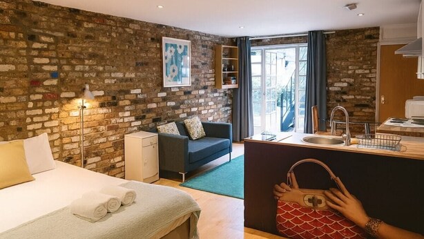 Quarto de hotel com parede de tijolos, cama com lençóis brancos, escrivaninha com pia e uma janela grande.