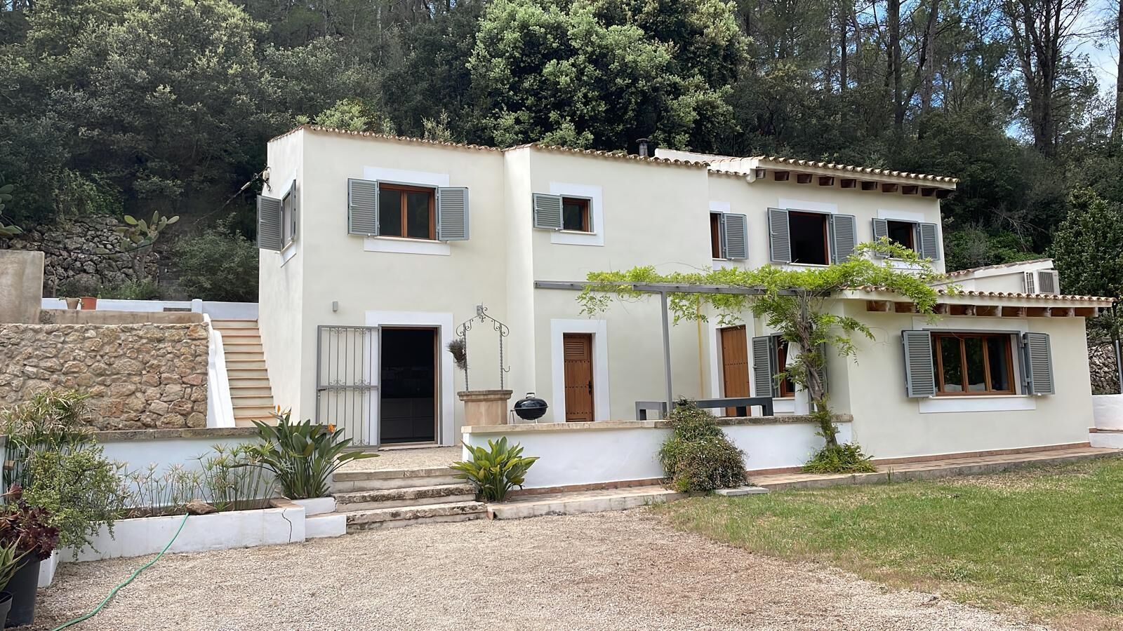Uma casa de dois andares com uma parede de pedra, uma porta de madeira e uma pequena área de jardim com plantas e uma churrasqueira.