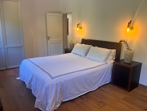 Una cama bien hecha con sábanas blancas y un cabecero oscuro, flanqueada por mesitas de noche con lámparas y un jarrón.