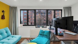 A room with a blue sofa, a teal chair, a yellow ottoman, a desk with a computer, a TV, and a window with a view of buildings. - Stylish 4 Bedroom 2 Bath House with Canal Views (London)