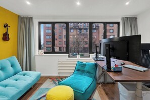 Ein Zimmer mit einem blauen Sofa, einem türkisfarbenen Sessel, einem gelben Hocker, einem Schreibtisch mit einem Computer, einem Fernseher und einem Fenster mit Blick auf Gebäude.