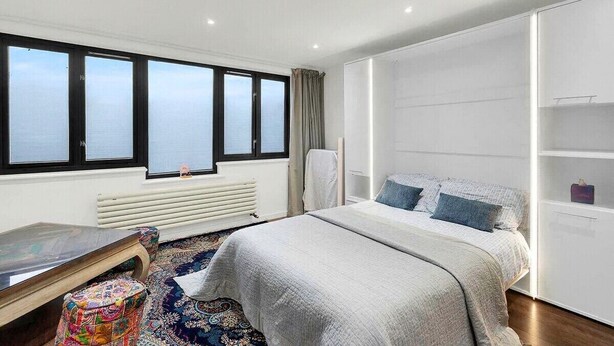 A modern hotel room with a large bed, a desk, a colorful ottoman, and a view of the sea through large windows. - Stylish 4 Bedroom 2 Bath House with Canal Views (London)