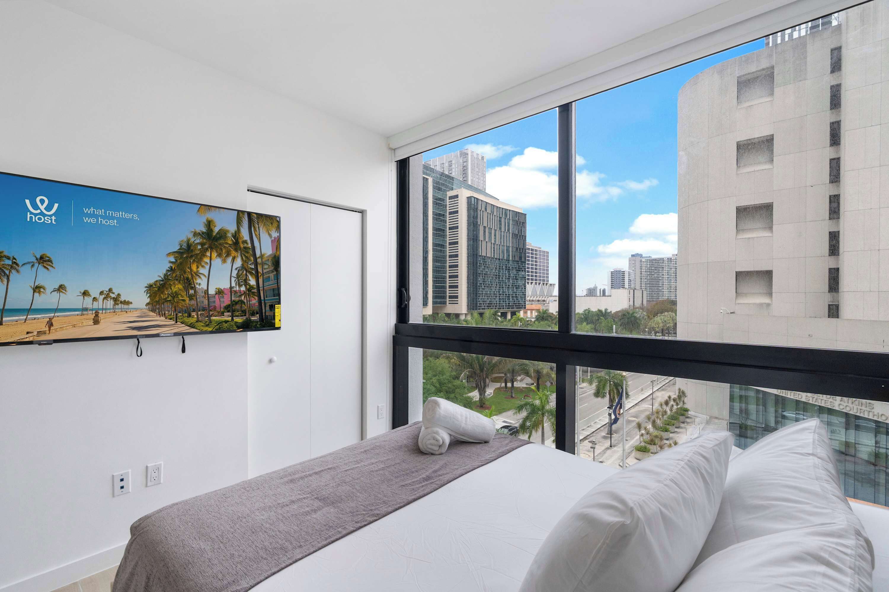 A hotel room with a bed, a window with a city view, a wall-mounted picture, and a power outlet.
