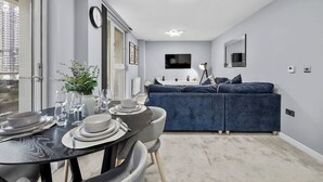 A modern living room with a sofa, a dining table set with plates and glasses, a TV mounted on the wall, and a ceiling light fixture. - 2-bedroom apartment near Canary Wharf (London)