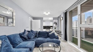 A modern living room with a blue sofa, a glass coffee table, and a city view through large windows. - 2-bedroom apartment near Canary Wharf (London)