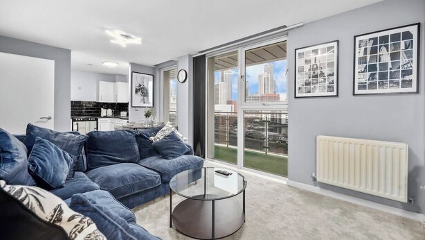 A modern living room with a sofa, coffee table, and a view of the city through large windows. - 2-bedroom apartment near Canary Wharf (London)