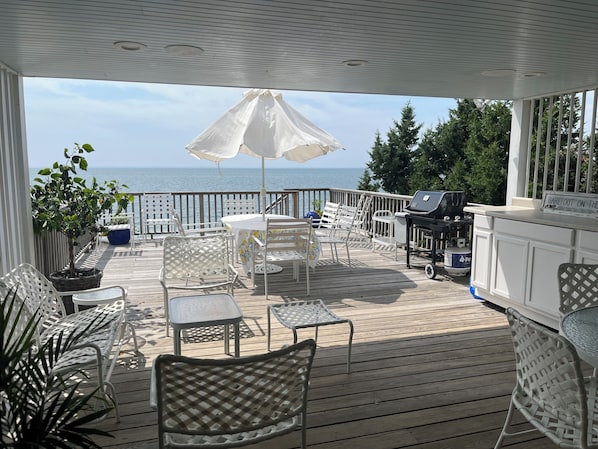 A covered outdoor area with a view of the sea, featuring a grill, tables, and chairs.