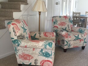 Une pièce avec un escalier, deux fauteuils recouverts d’un tissu à motifs marins, une lampe et une table à manger avec des chaises.