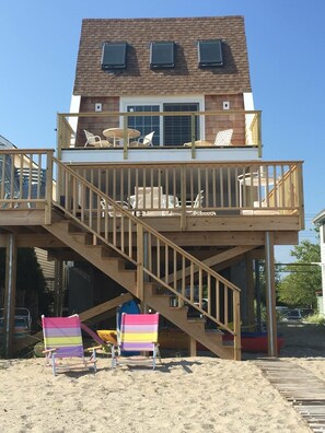 Una casa de dos pisos con terraza de madera, escaleras y mobiliario exterior.