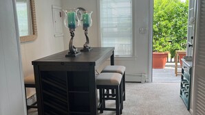Ein Zimmer mit einem dunklen Holztisch, zwei Stühlen und einer Bank. Zwei brennende Kerzen stehen auf dem Tisch. Ein Fenster mit Jalousien ist sichtbar, und eine Tür führt in einen Außenbereich mit Topfpflanzen.