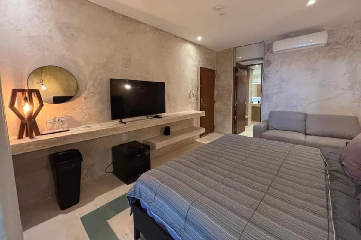 A modern bedroom with a large bed, a flat-screen TV mounted on a marble wall, a small seating area with a sofa, and a trash bin.