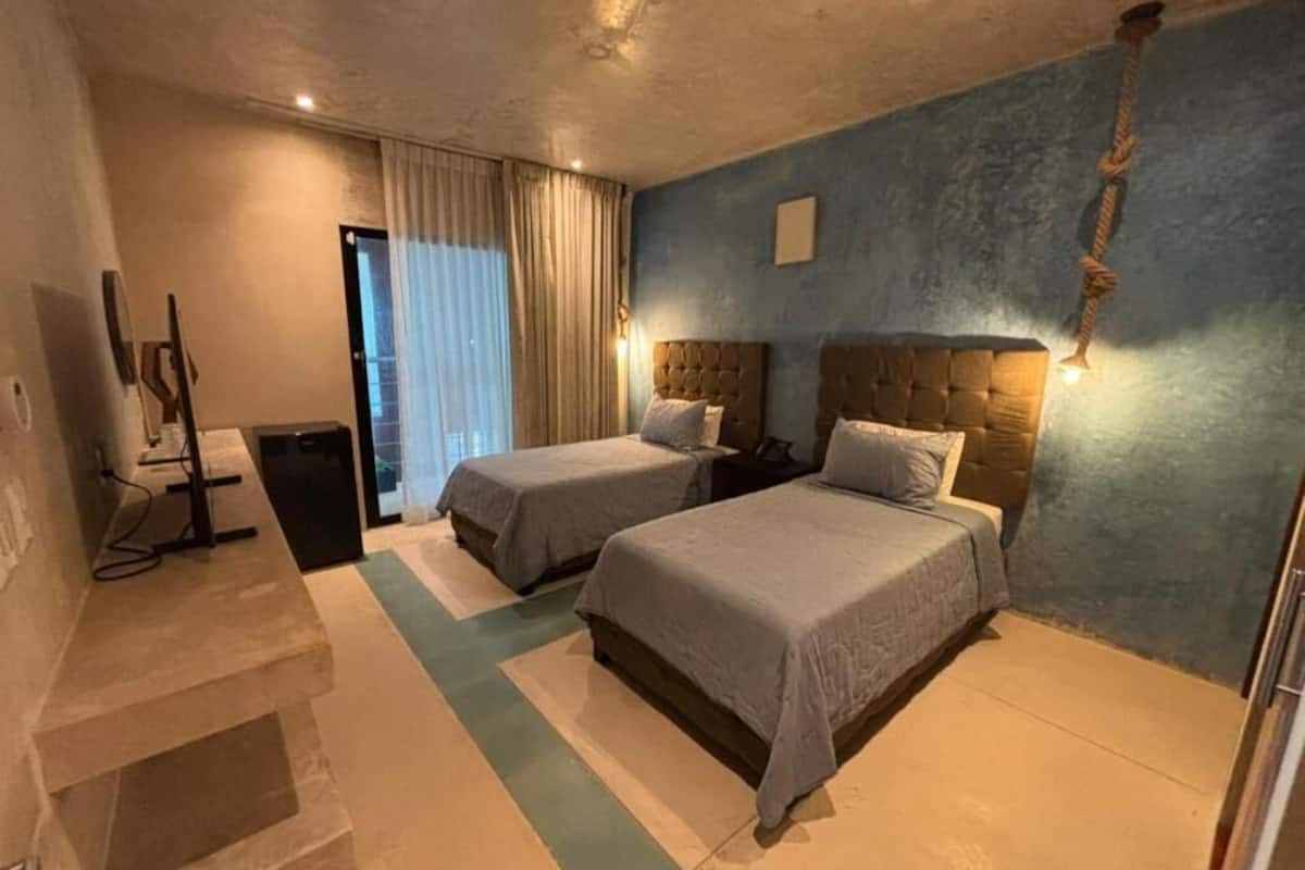 A hotel room with two beds, a desk, a trash bin, and a window with curtains.