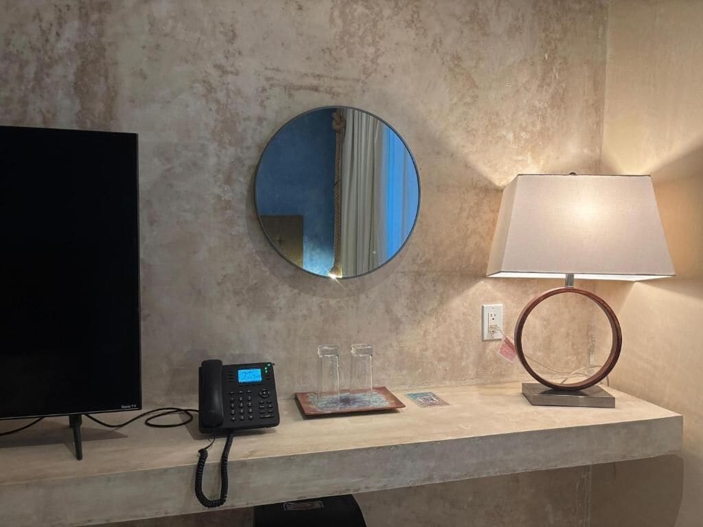 Zona de recepción de un hotel con una televisión de pantalla plana, un teléfono digital, un espejo redondo, una lámpara de mesa y un pequeño objeto decorativo.