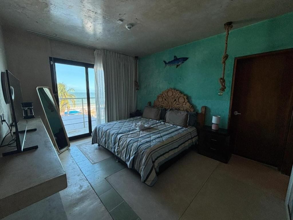 A bedroom with a bed, a desk, a chair, a window with a view of the beach, and a door.