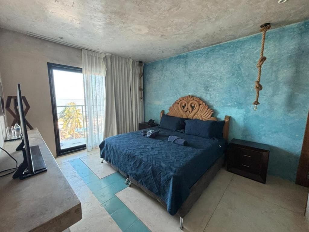 Une chambre avec un grand lit, une tête de lit en bois, une table de chevet, un bureau avec un ordinateur et une vue à travers une porte coulissante.