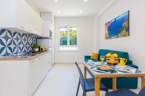 A compact kitchen with a window, a dining area with a table set for breakfast, and a blue sofa. - Dolce Vita Apartments - Capri with Private Terrace, Wi-Fi and Air Conditioning (Sorrento)