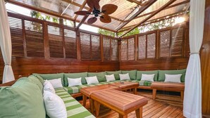 Un espace extérieur abrité, aménagé avec du mobilier en bois et doté d’un ventilateur de plafond.