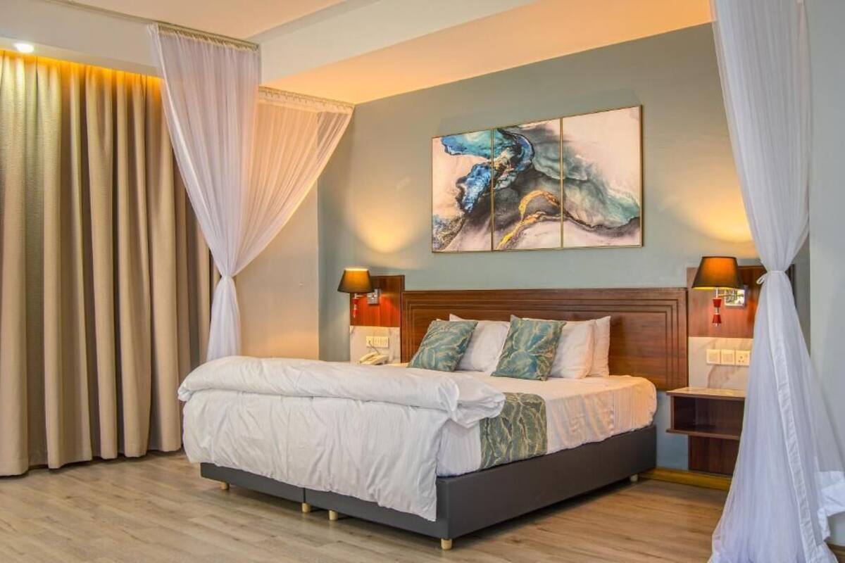 Een slaapkamer met een bed, gordijnen, een schilderij, bedlampjes en een nachtkastje.