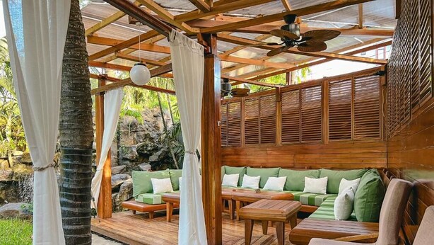 Une terrasse extérieure aménagée avec du mobilier en bois, notamment des canapés et une table, sous une pergola en bois dotée d’un ventilateur de plafond.