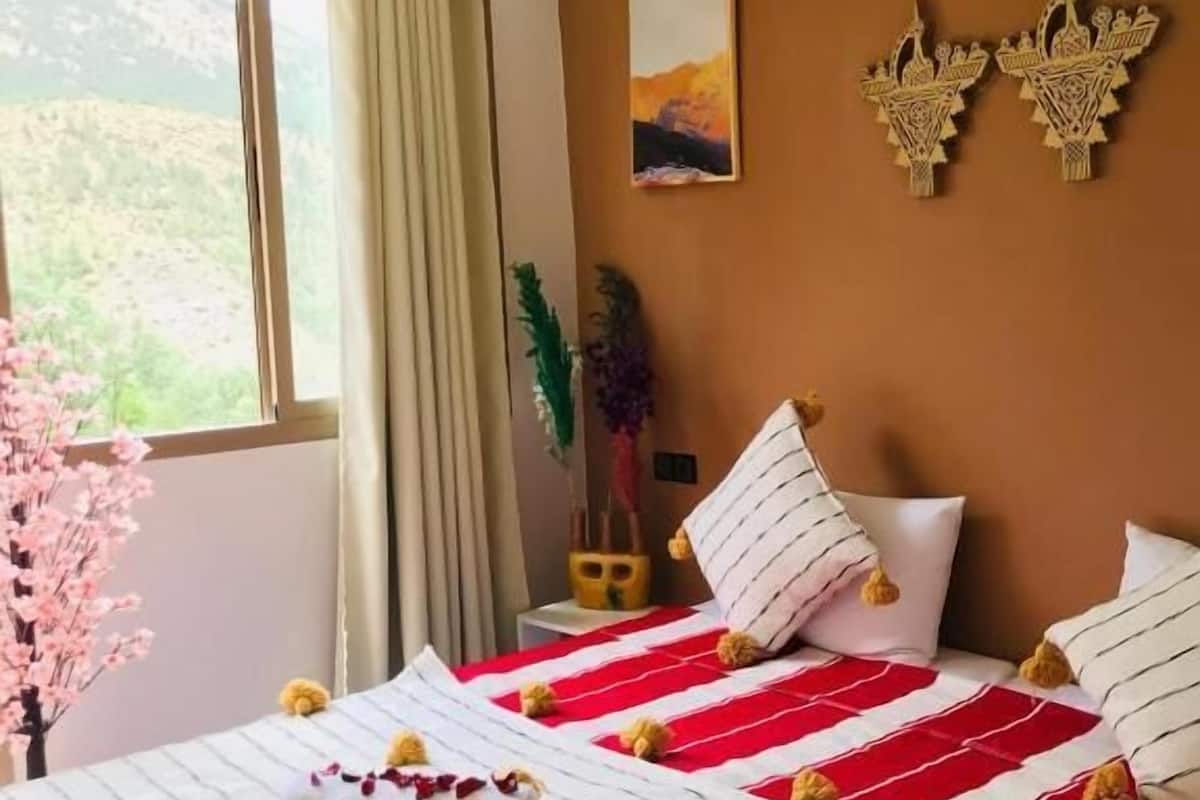 A bedroom with a bed, pillows, a window, curtains, a wall-mounted artwork, and a decorative wall piece.