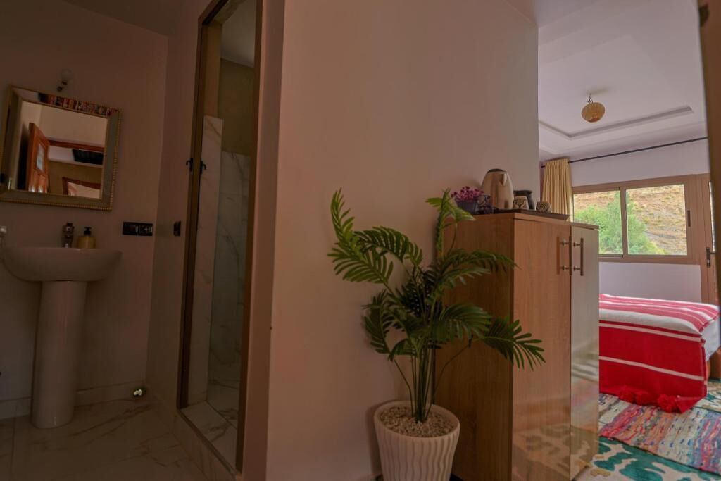 Une salle de bain avec un lavabo, un miroir et une douche. On y trouve une plante en pot et un meuble en bois, et on peut apercevoir un lit à travers une porte ouverte.