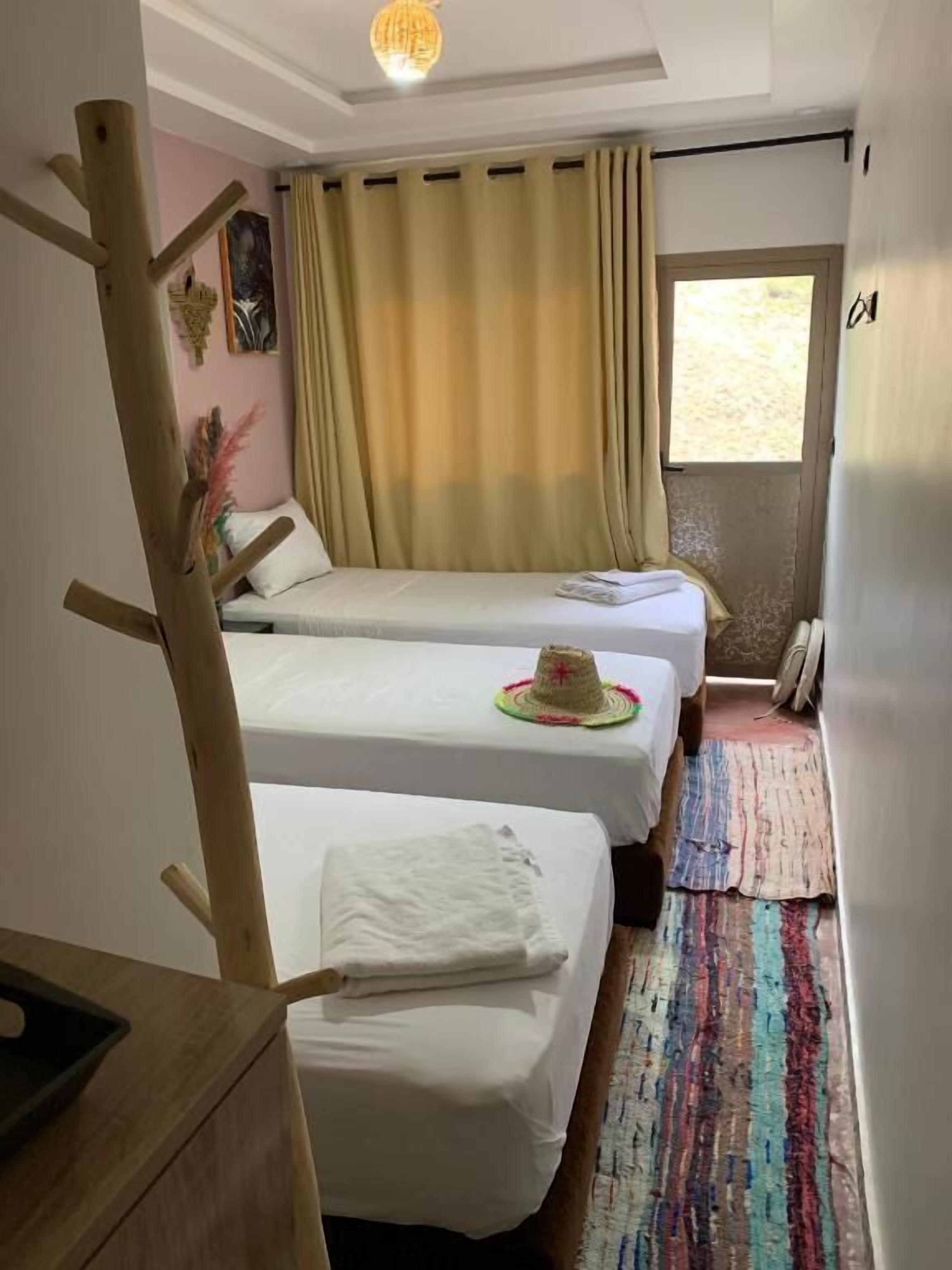 Una habitación pequeña y acogedora con dos camas individuales, una silla de madera, una mesita y una ventana con cortinas.