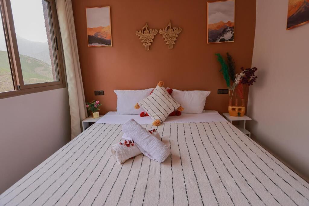 A neatly made bed with white linens and striped pillows, a small bedside table with a vase of flowers, and a window with a view of the outdoors.