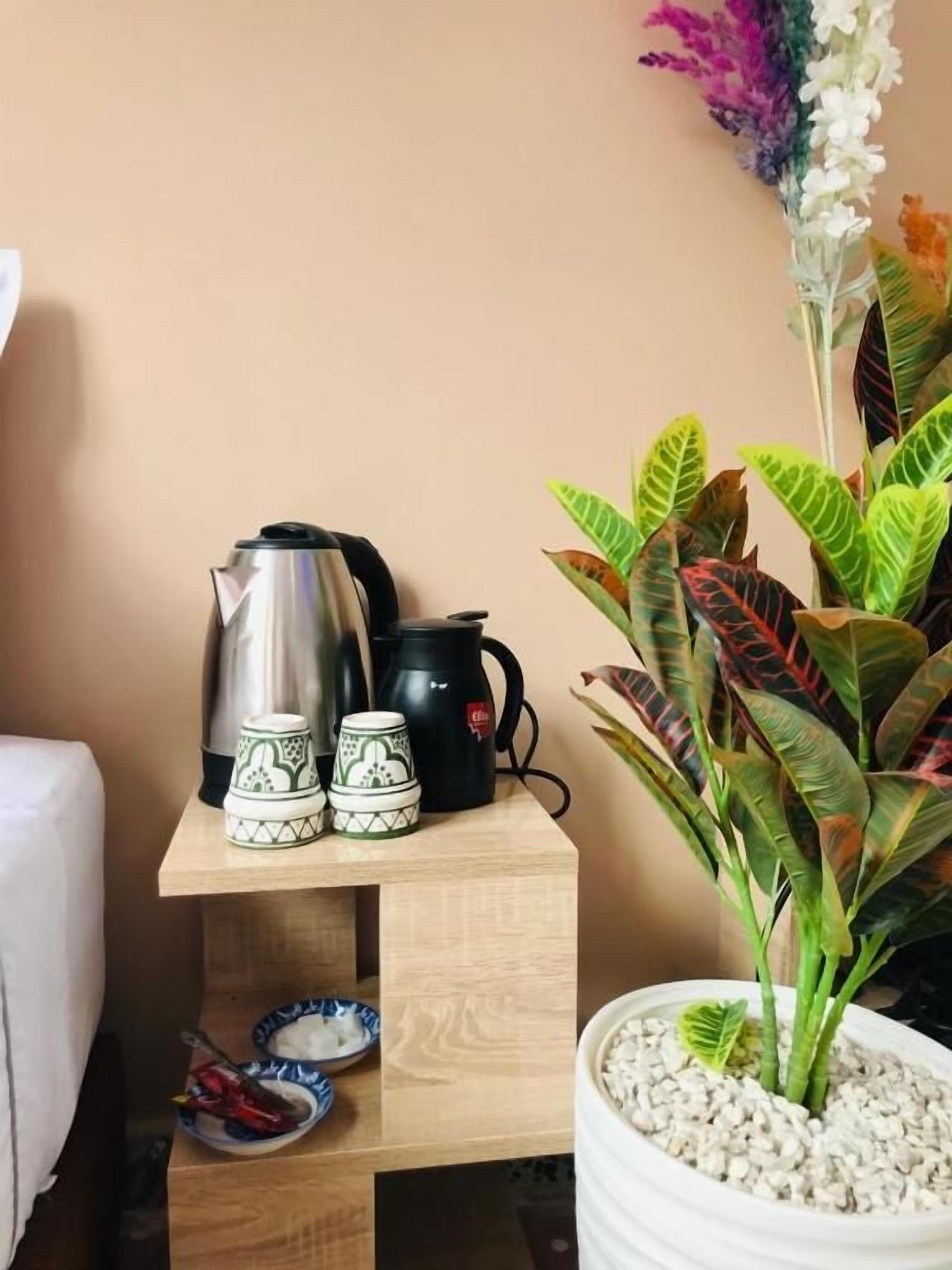 A wooden shelf with a kettle, a black coffee pot, and two cups with green patterns. There is a potted plant with broad leaves and a white pot filled with pebbles.