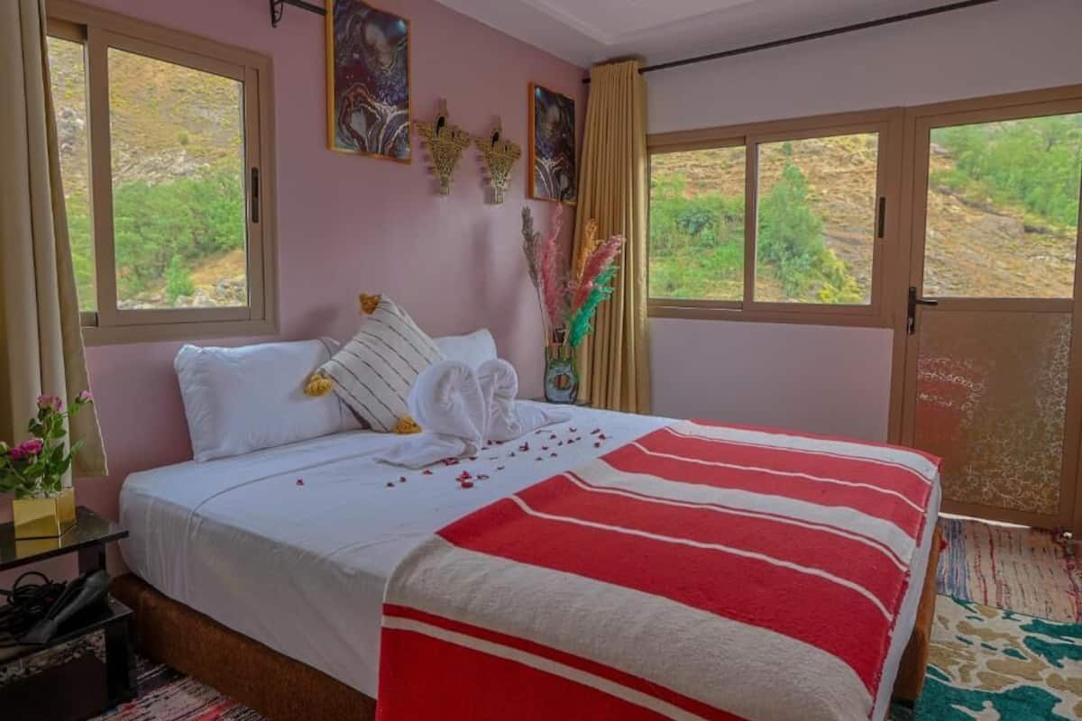 A bedroom with a bed, a window, a door, and a view of greenery outside.