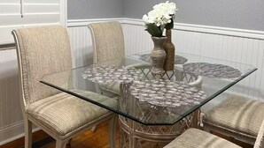 A dining area with a glass-top table surrounded by wicker chairs, a vase with white flowers, and wall decorations including a map and signs. - Family Friendly home down from the lake, just minutes to a dock and downtown! (Guntersville)