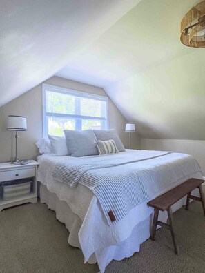 A bedroom with a sloped ceiling, a large window, a bed with white bedding, two bedside lamps, and a wooden bench. - Lakefront sunsets, dock fishing & cozy fire · 20 min to Mad River (Belle Center)