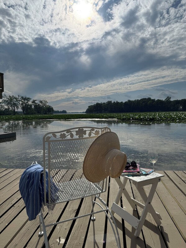 A deck chair with a hat and a glass of wine on a wooden deck overlooking a calm body of water with lily pads and trees in the background.