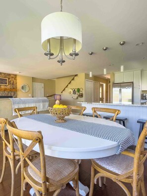 A dining area with a round table, wicker chairs, a chandelier, and a kitchen in the background.