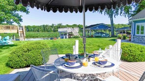 An outdoor patio with a round table set for breakfast, surrounded by chairs, and a covered area with a view of a lake and a house. - Lakefront sunsets, dock fishing & cozy fire · 20 min to Mad River (Belle Center)